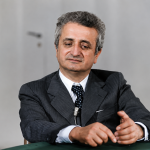 Ritratto storico di Vittorino Colombo durante un'intervista, seduto al tavolo con microfono, in un momento della sua attività politica e istituzionale.