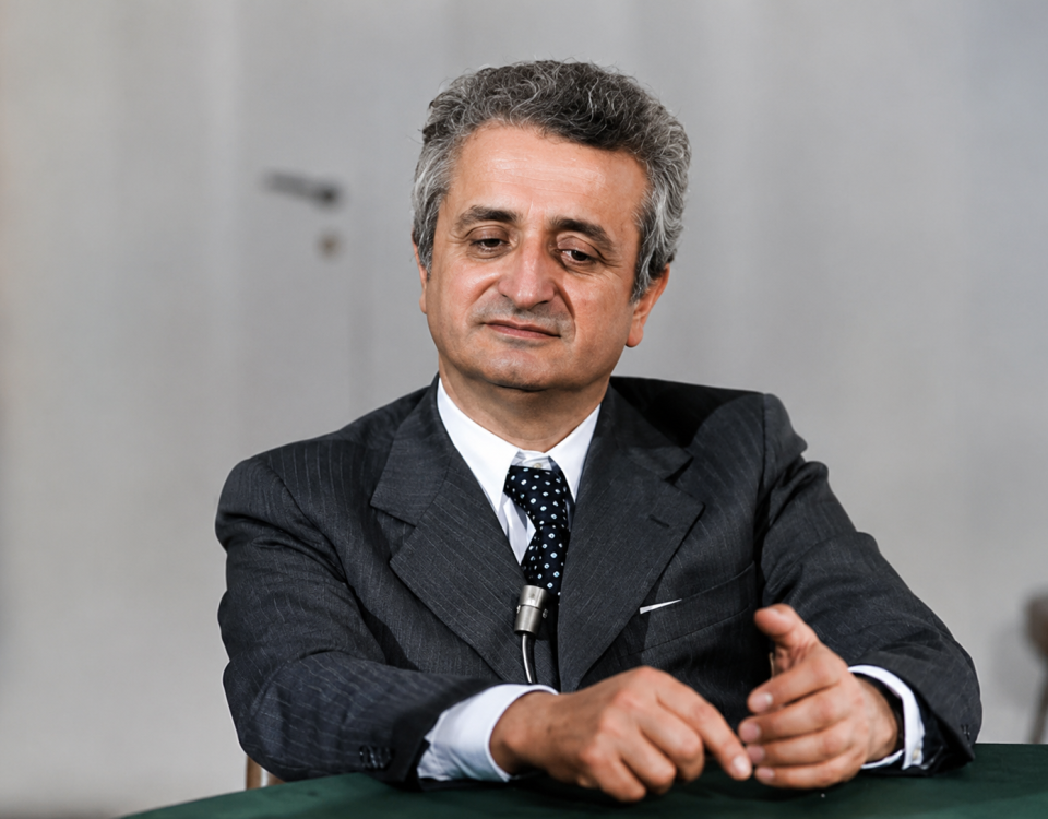 Ritratto storico di Vittorino Colombo durante un'intervista, seduto al tavolo con microfono, in un momento della sua attività politica e istituzionale.
