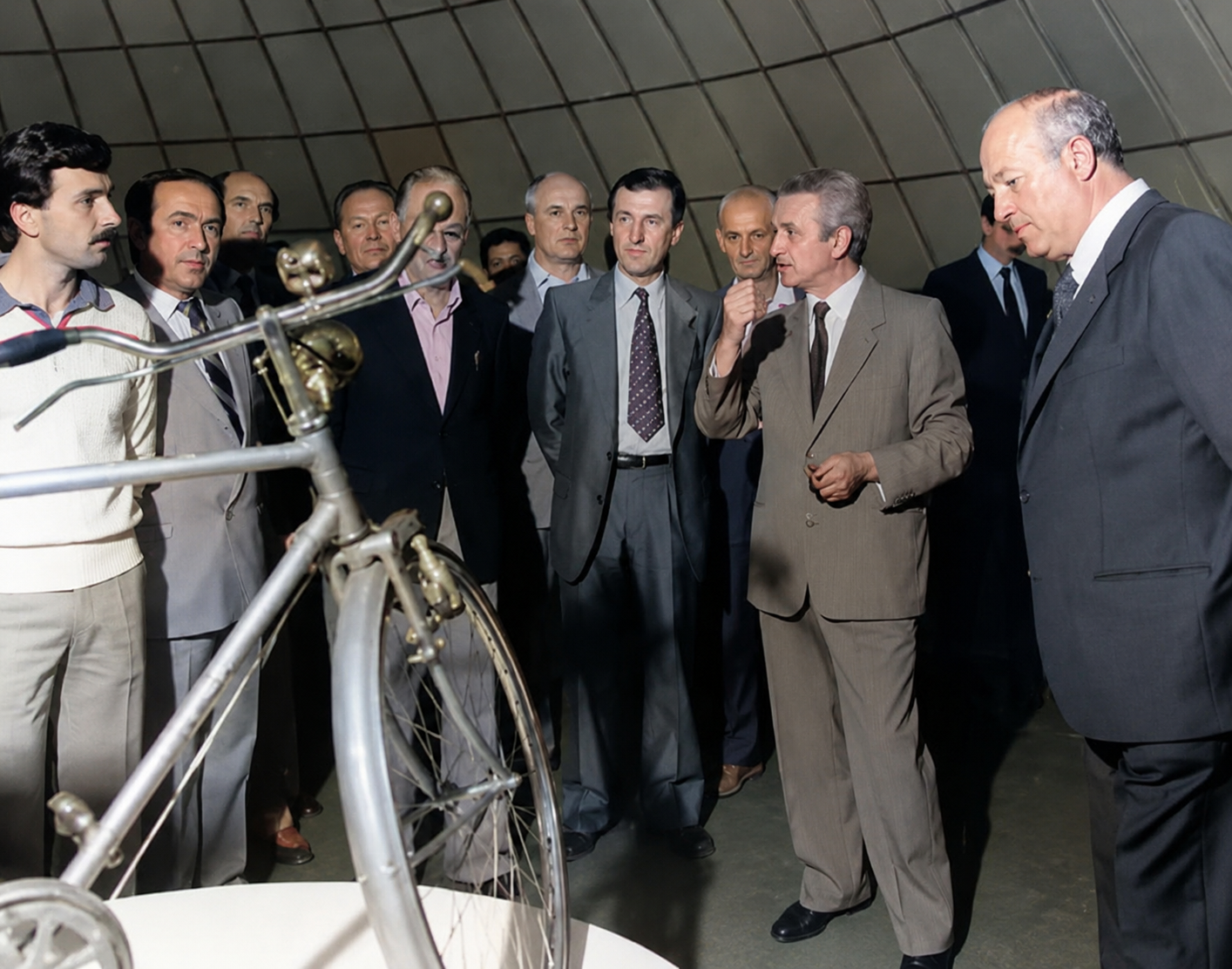 Vittorino Colombo osserva una bicicletta storica durante una visita istituzionale insieme a una delegazione di rappresentanti politici e culturali.